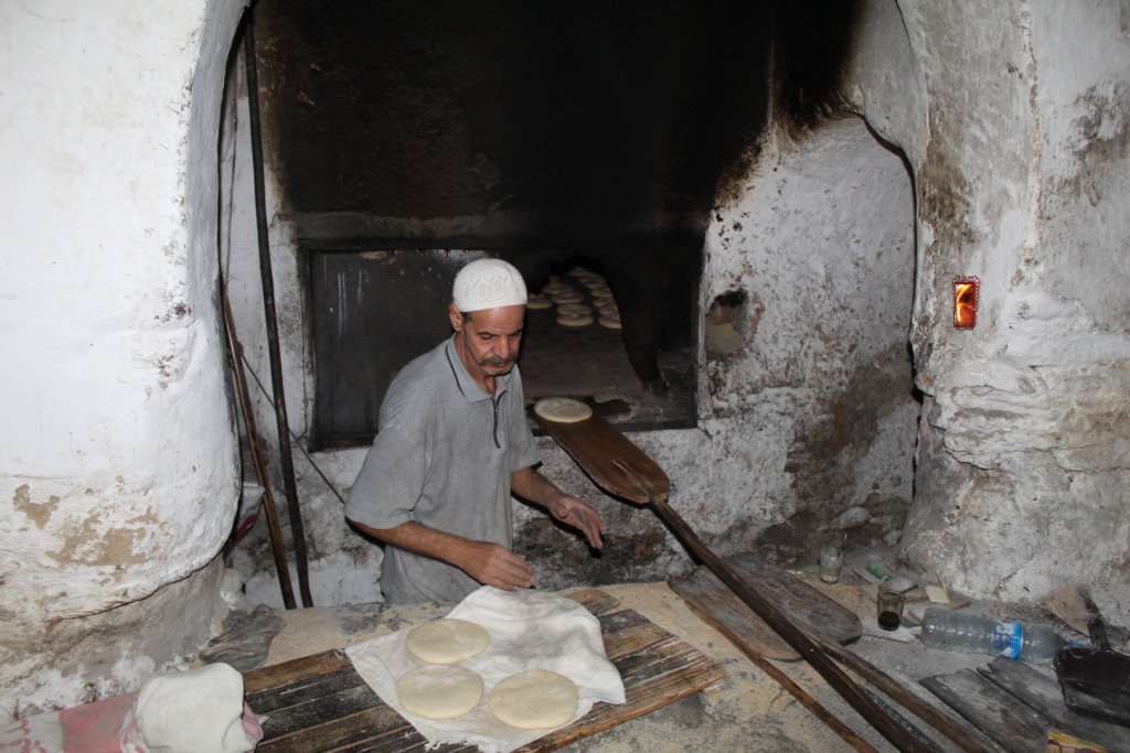 This photo shows a baker in a sunken oven loading bread with long wooden paddles